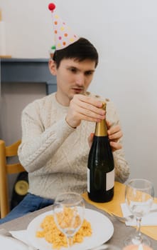 One young handsome Caucasian brunette guy with a cone hat on his head opens a bottle of champagne while sitting at the table and celebrating his birthday, close-up side view.
