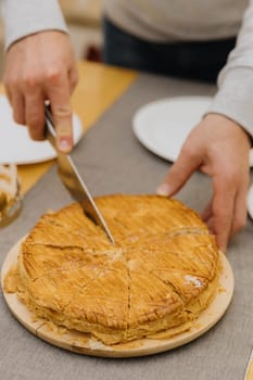 One Caucasian young blurred man cuts a royal galette in a plate on a wooden table with a knife, standing at the table, close-up side view with selective focus.