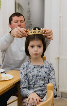 One young handsome Caucasian man puts a golden paper crown on his little girl daughter after eating a royal galette and finding a gift, while sitting at the table in the kitchen, close-up side view.