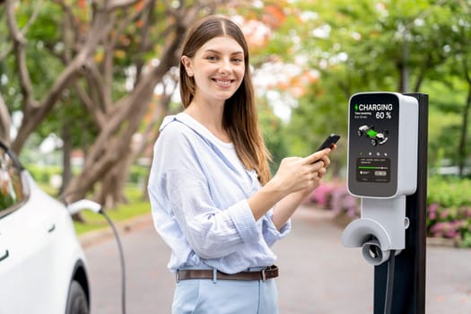 Young woman using smartphone online banking application to pay for electric car battery charging from EV charging station during autumn vacation holiday trip at national park or autumnal forest. Exalt