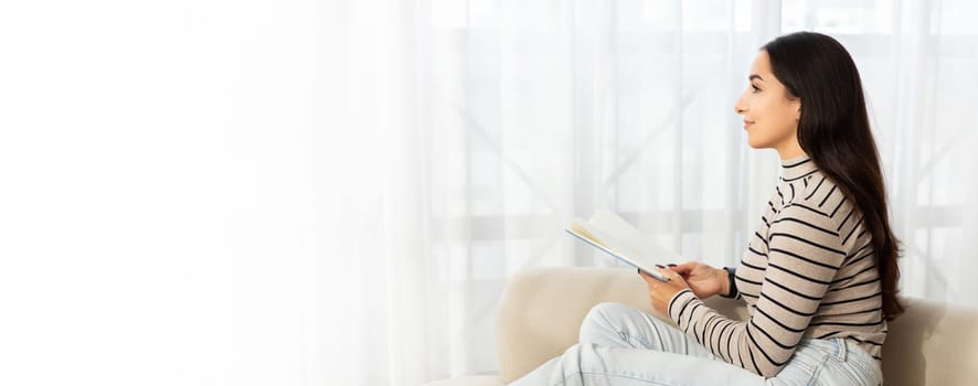 Smiling pensive calm young caucasian lady read book, planning work, sit at sofa, dream in light living room interior. Free time, study, education, hobby and lifestyle cozy at home