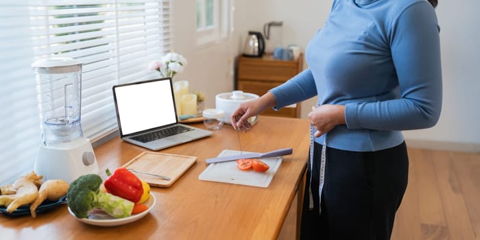 Cheerful Fat woman cooking in kitchen showing laptop mock up blank white screen in home.