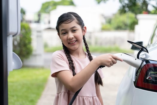 Happy little young girl learn about eco-friendly and energy sustainability as she recharge electric vehicle from home EV charging station. EV car and sustainable future generation concept. Synchronos