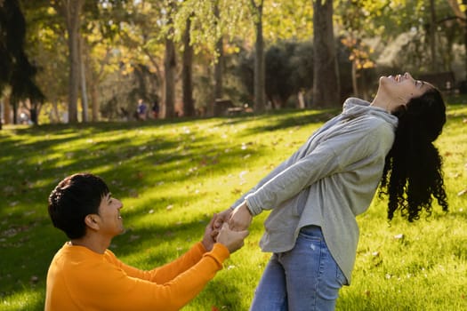 Happy young latin couple enjoy time together on a public park. Boyfriend holding girlfriend hands asking for marriage.