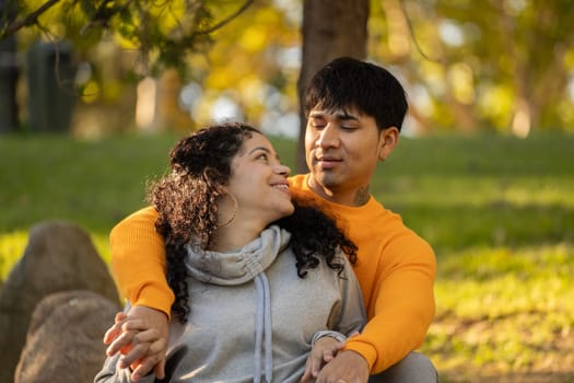 Latin couple hugging and flirting in an urban park sitting in the grass and looking each other.