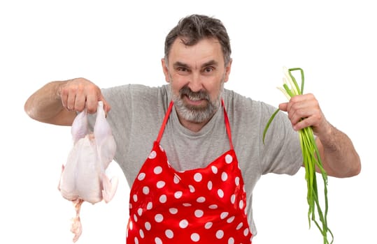 An elderly bearded male cook in a red polka-dot apron holds a raw chicken and a bunch of onions on a white background. Cook with chicken.