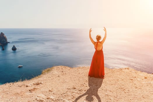 Side view a Young beautiful sensual woman in a red long dress posing on a rock high above the sea during sunrise. Girl on the nature on blue sky background. Fashion photo.