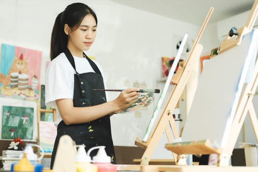 Young female artist sketches or paints her drawing on canvas in a studio workshop. A teenage girl who likes art and drawing is taking time to create her watercolors on canvas with great intention. Artist studio interior. Drawing supplies, oil paints, artist brushes, canvas, frame. Workshop or art class. Creative concept..