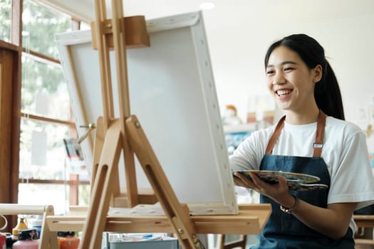 Young female artist sketches or paints her drawing on canvas in a studio workshop. A teenage girl who likes art and drawing is taking time to create her watercolors on canvas with great intention. Artist studio interior. Drawing supplies, oil paints, artist brushes, canvas, frame. Workshop or art class. Creative concept..