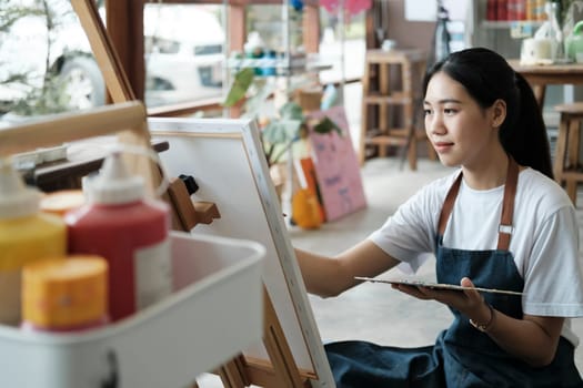 Young female artist sketches or paints her drawing on canvas in a studio workshop. A teenage girl who likes art and drawing is taking time to create her watercolors on canvas with great intention. Artist studio interior. Drawing supplies, oil paints, artist brushes, canvas, frame. Workshop or art class. Creative concept..