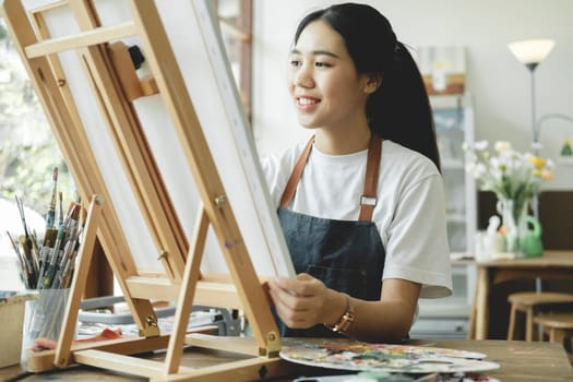 Young female artist sketches or paints her drawing on canvas in a studio workshop. A teenage girl who likes art and drawing is taking time to create her watercolors on canvas with great intention. Artist studio interior. Drawing supplies, oil paints, artist brushes, canvas, frame. Workshop or art class. Creative concept..