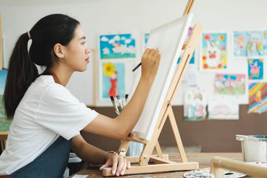 Young female artist sketches or paints her drawing on canvas in a studio workshop. A teenage girl who likes art and drawing is taking time to create her watercolors on canvas with great intention. Artist studio interior. Drawing supplies, oil paints, artist brushes, canvas, frame. Workshop or art class. Creative concept..