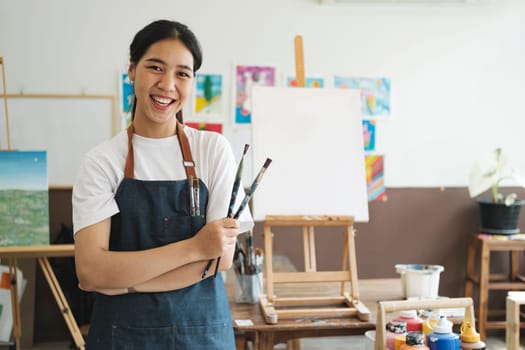 The young artist female is wearing an apron smeared with paint clutching many brushes and paintbrushes stnading and smiling to the camera in her studio workshop or art gallery. Hobby and lifestyle concept.