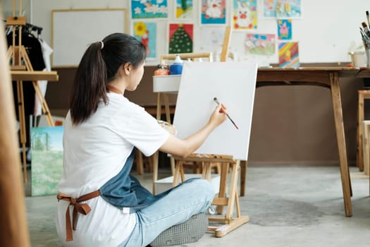 Young female artist sketches or paints her drawing on canvas in a studio workshop. A teenage girl who likes art and drawing is taking time to create her watercolors on canvas with great intention. Artist studio interior. Drawing supplies, oil paints, artist brushes, canvas, frame. Workshop or art class. Creative concept..