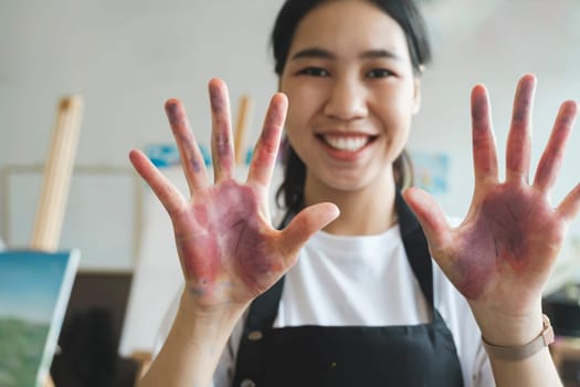 Creative portrait, woman hands with paint on happy art school student and oil painting inspiration for Indian girl. Fun learning color theory and young artist creativity in university workshop studio. The artist creates a frame with her hands soiled with watercolor paints.