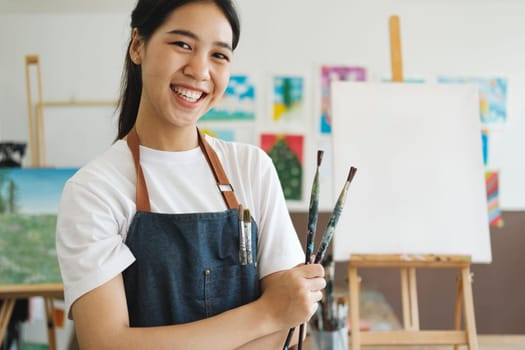 The young artist female is wearing an apron smeared with paint clutching many brushes and paintbrushes stnading and smiling to the camera in her studio workshop or art gallery. Hobby and lifestyle concept.