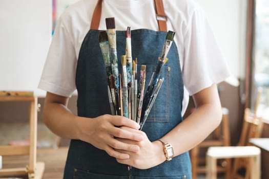 A close-up of the artist hands wearing an apron smeared with paint. Clutching many brushes and paintbrushes. Hobby and lifestyle concept.