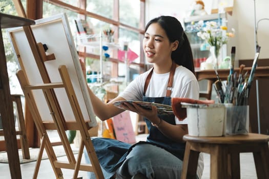 Young female artist sketches or paints her drawing on canvas in a studio workshop. A teenage girl who likes art and drawing is taking time to create her watercolors on canvas with great intention. Artist studio interior. Drawing supplies, oil paints, artist brushes, canvas, frame. Workshop or art class. Creative concept..