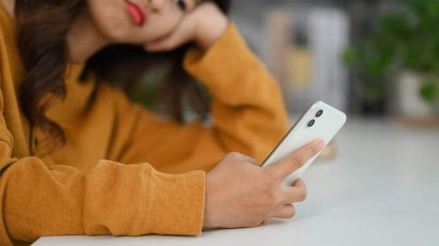 Bored asian woman leaning on hand using smartphone at home, waiting on slow wifi connection