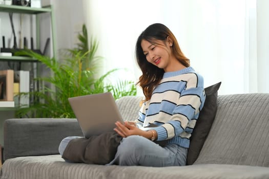 Relaxed young Asian woman sitting on comfortable couch and using laptop.