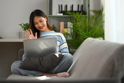 Happy young Asian woman waving hand video calling via laptop, sitting on couch at home.