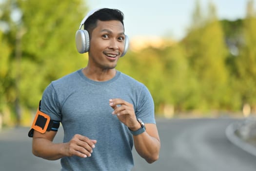 Happy millennial man listening to music in headphones, jogging in the park on a sunny day.