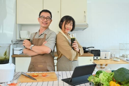 Portrait retirement senior couple posing and smiling at camera while standing in kitchen.