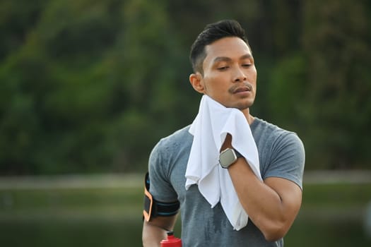 Young muscular man resting and wiping sweat with a towel after morning cardio workout in park.