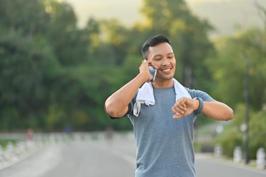 Smiling sportsman having pleasant phone conversation and checking time on watch while exercise in the park.