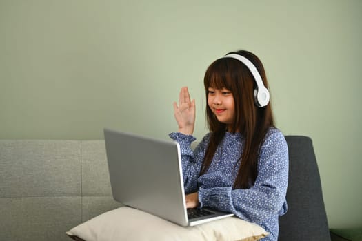 Smiling Asian schoolgirl studying online at home looking at laptop and waving hand. Online education concept