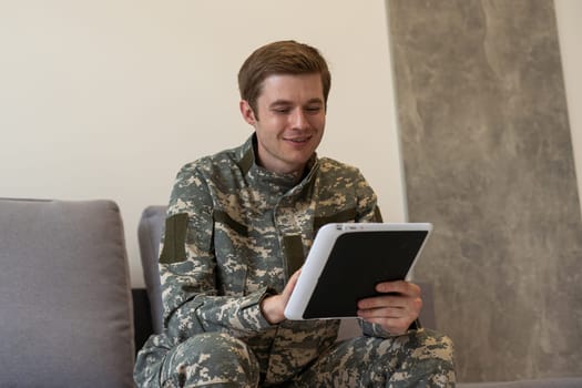 Smiling millennial man in camouflage uniform holding modern digital tablet, chatting with family, posing on white studio background, copy space. Modern technologies and military personnel concept