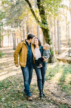 Dad almost kisses mom with a little girl in her arms while walking in the forest. High quality photo