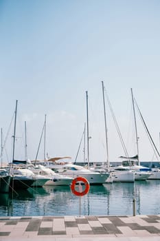 Red lifebuoy stands on a stand on the pier against the background of moored yachts. High quality photo