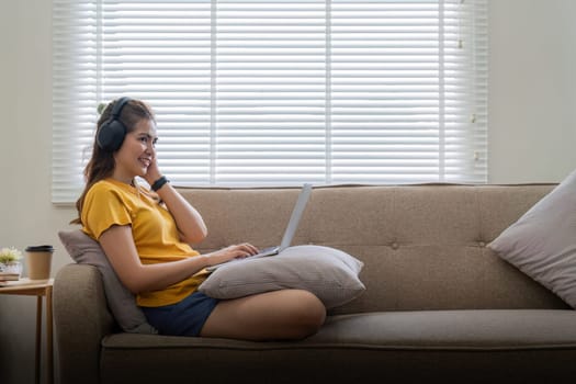 Happy woman wearing headphones watching and listening video on laptop and listening music sitting on a couch at home.