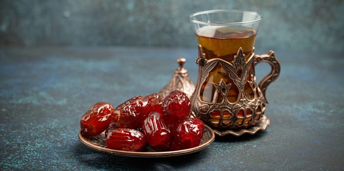 Breaking fasting with dried dates during Ramadan Kareem, Iftar meal with dates and Arab tea in traditional glass, angle view on rustic blue background. Muslim feast.