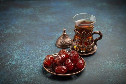 Breaking fasting with dried dates during Ramadan Kareem, Iftar meal with dates and Arab tea in traditional glass, angle view on rustic blue background. Muslim feast.