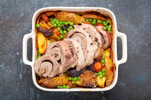 Rolled sliced pork roasted in white casserole dish with potatoes, vegetables and herbs on blue dark concrete rustic background top view. Baked pork roll with vegetables for dinner.