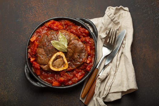 Traditional Italian dish Ossobuco all Milanese made with cut veal shank meat with vegetable tomato sauce served in black casserole pan top view on rustic brown background.