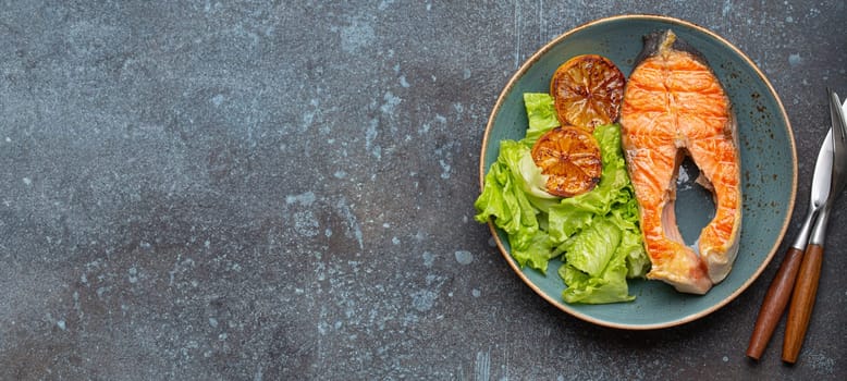 Grilled fish salmon steak and green salad with lemon on ceramic plate on rustic blue stone background top view, balanced diet or healthy nutrition meal with salmon and veggies.