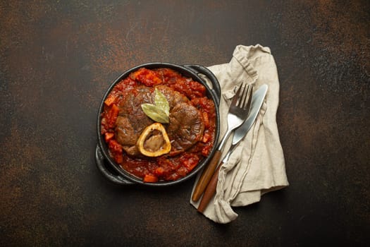 Traditional Italian dish Ossobuco all Milanese made with cut veal shank meat with vegetable tomato sauce served in black casserole pan top view on rustic brown background.