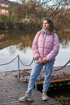 A young woman standing at the shore looking at the river in autumn sunny day. Street view, copy space for text, travel photo. Happy tourist woman on the bank of the river in autumn in warm clothes. Tourists enjoy their vacation, winter season. Romantic look and travel concept. A joyful mood in a Caucasian girl. Winter Wonderland: Enchanting Girl by the Riverside in Autumn.