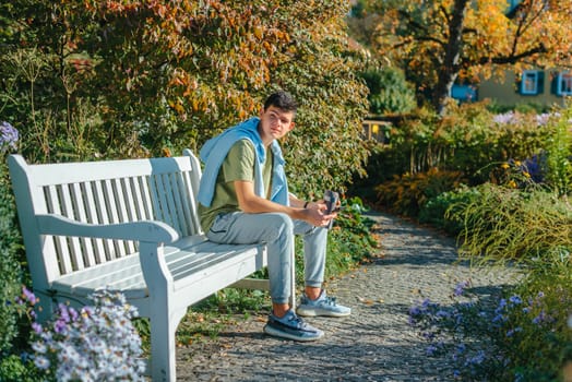 a teenager sits on a bench in the park drinks coffee from a thermo mug and looks into a phone. Portrait of handsome cheerful guy sitting on bench fresh air using device browsing media smm drinking latte urban outside outdoor.