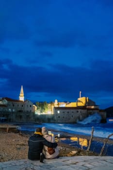 Couple sits embracing on the seashore and looks at the illuminated ancient town. Back view. High quality photo