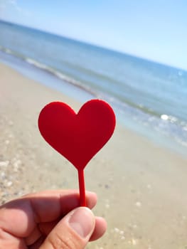 Person holding in fingers hand stick in shape red heart on background sea and sea waves, blue sky in seashore on sunny summer day close-up. Concept love romance amour St Valentines Day