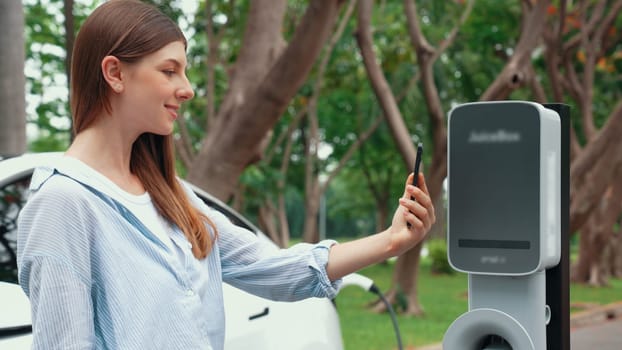 Woman using smartphone online banking application to pay for electric car battery charging from EV charging station during vacation holiday road trip at national park or summer forest. Exalt