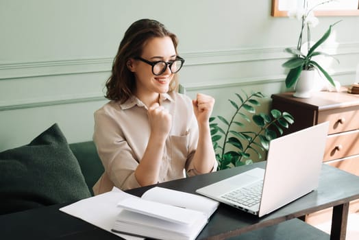 Victory and Joy. Smiling Woman Gesturing in Successful Laptop Video Chat. Triumphant Video Call. Joyful Woman in Glasses Beams as She Communicates on Laptop, Signaling Victory and Success.