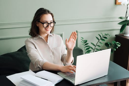 Happy Young Woman in Glasses Engaged in Joyful Laptop Video Chat. Energetic Video Chat: Joyful Woman in Glasses Engages in a Laptop Call, Waving Cheerfully.