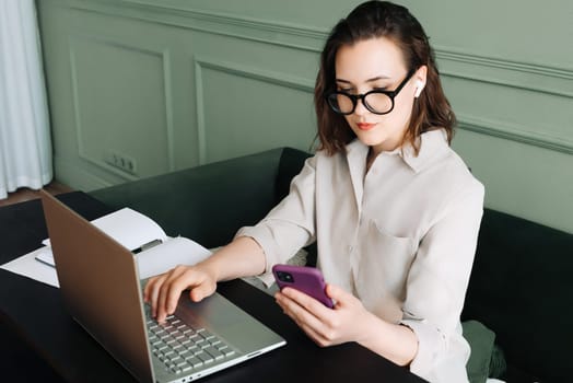 Productive Conversations. Woman Balancing Laptop and Smartphone in Video Call. Multitasking Joy. Busy Woman in Glasses Juggles Video Call on Laptop and Smartphone with a Smile.