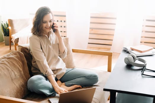 Multitasking in Style. Busy Young Woman on Sofa with Smartphone and Laptop. Sitting on the sofa in her living room, making calls on her smartphone and typing on her laptop, happily busy young woman