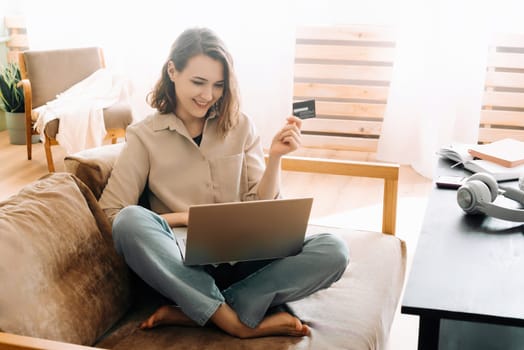 A cheerful woman typing on a laptop, using a credit card, shopping online in a room sitting on the couch. Happy Shopping Moment. Woman Using Laptop and Credit Card on Sofa.